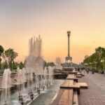 A wide-angle view of a monumental neoclassical building in Dushanbe, Tajikistan, featuring grand columns and a manicured park with a fountain in the foreground under a clear sunset.