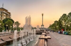 A wide-angle view of a monumental neoclassical building in Dushanbe, Tajikistan, featuring grand columns and a manicured park with a fountain in the foreground under a clear sunset.