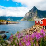 A traditional red wooden rorbu cabin on the water's edge in the Lofoten Islands, Norway, with steep mountains in the background.