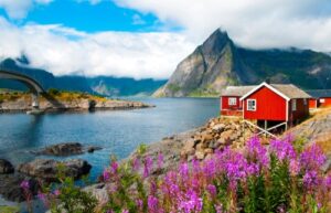 A traditional red wooden rorbu cabin on the water's edge in the Lofoten Islands, Norway, with steep mountains in the background.