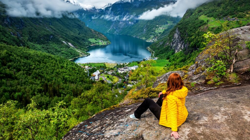 Travel blogger Sara Keelan sitting at a viewpoint overlooking Geirangerfjord’s steep cliffs and deep blue water in Norway.