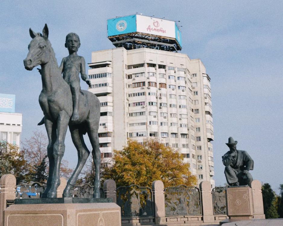 Statue of a child on horseback beside a seated figure with a tall apartment building in the background in Almaty, Kazakhstan.