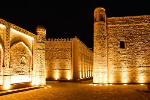 Illuminated brick fortress walls, towers, and an arched gateway in Khiva, Uzbekistan, at night.