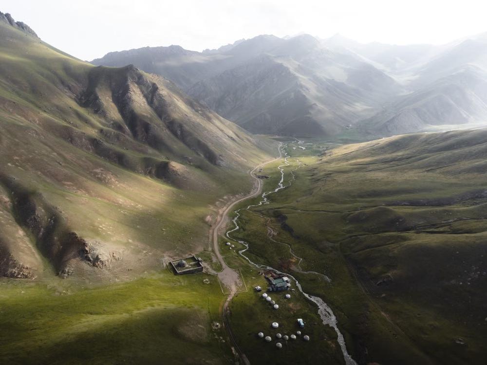 Wide mountain valley with a winding stream, dirt road, and scattered yurts in Kyrgyzstan.