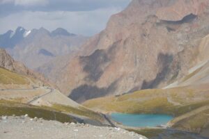 Mountain road winding beside a turquoise alpine lake beneath rugged peaks in Tajikistan.