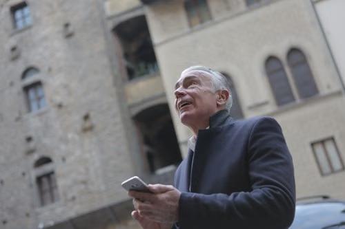 Older man holding a smartphone and looking up while standing among historic stone buildings.